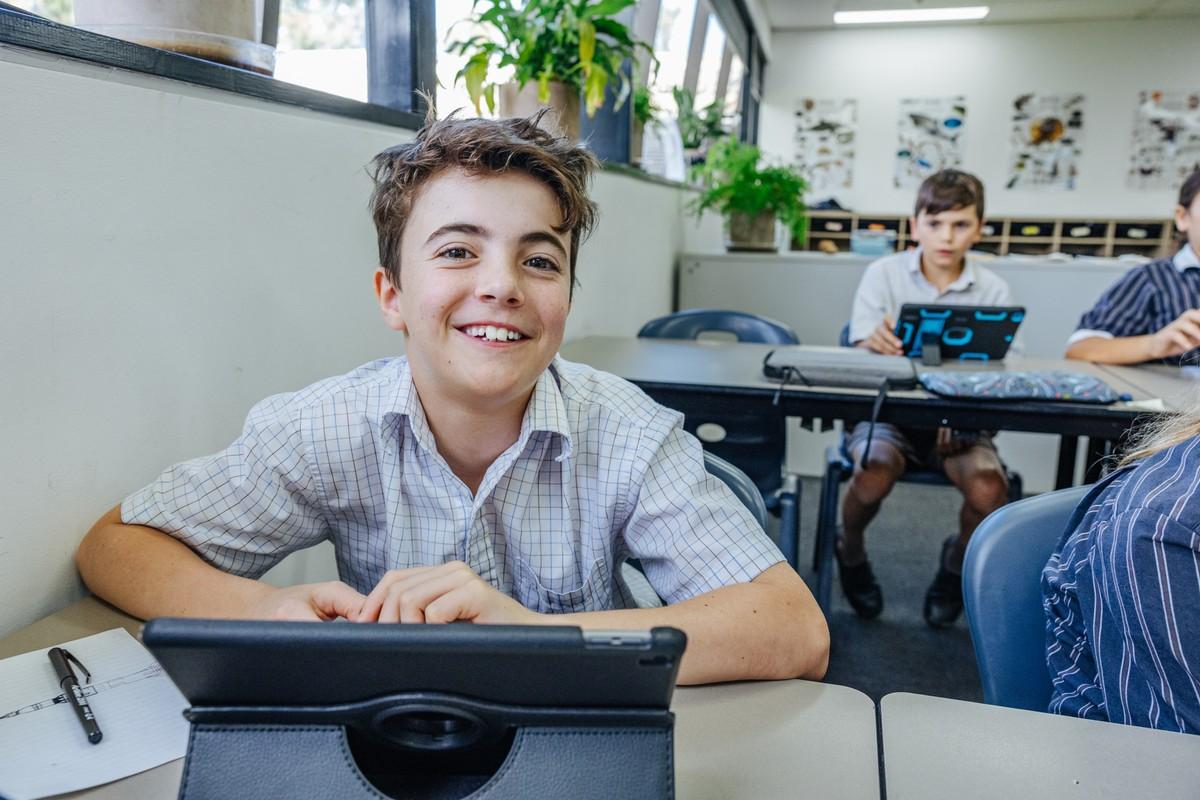 A student smiling at his desk with a tablet in a bright classroom, linking to booking a school tour.