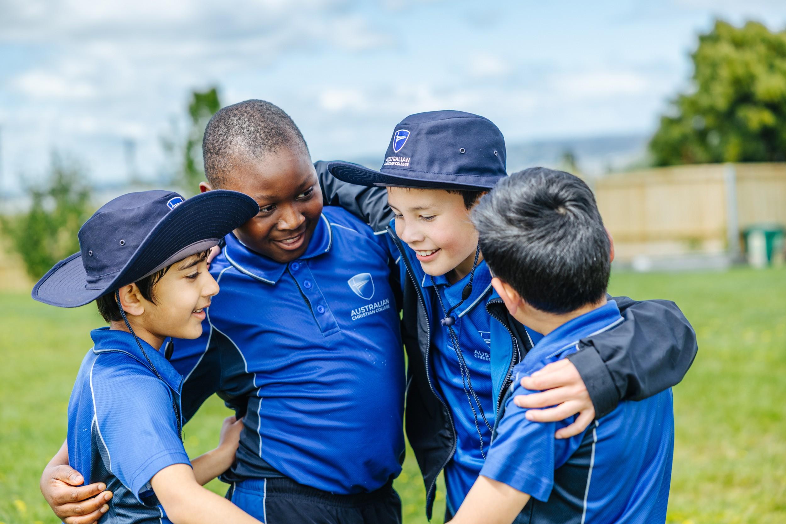 Four primary students in blue sports uniform and hats with arms around each other on the oval at ACC Bairnsdale.