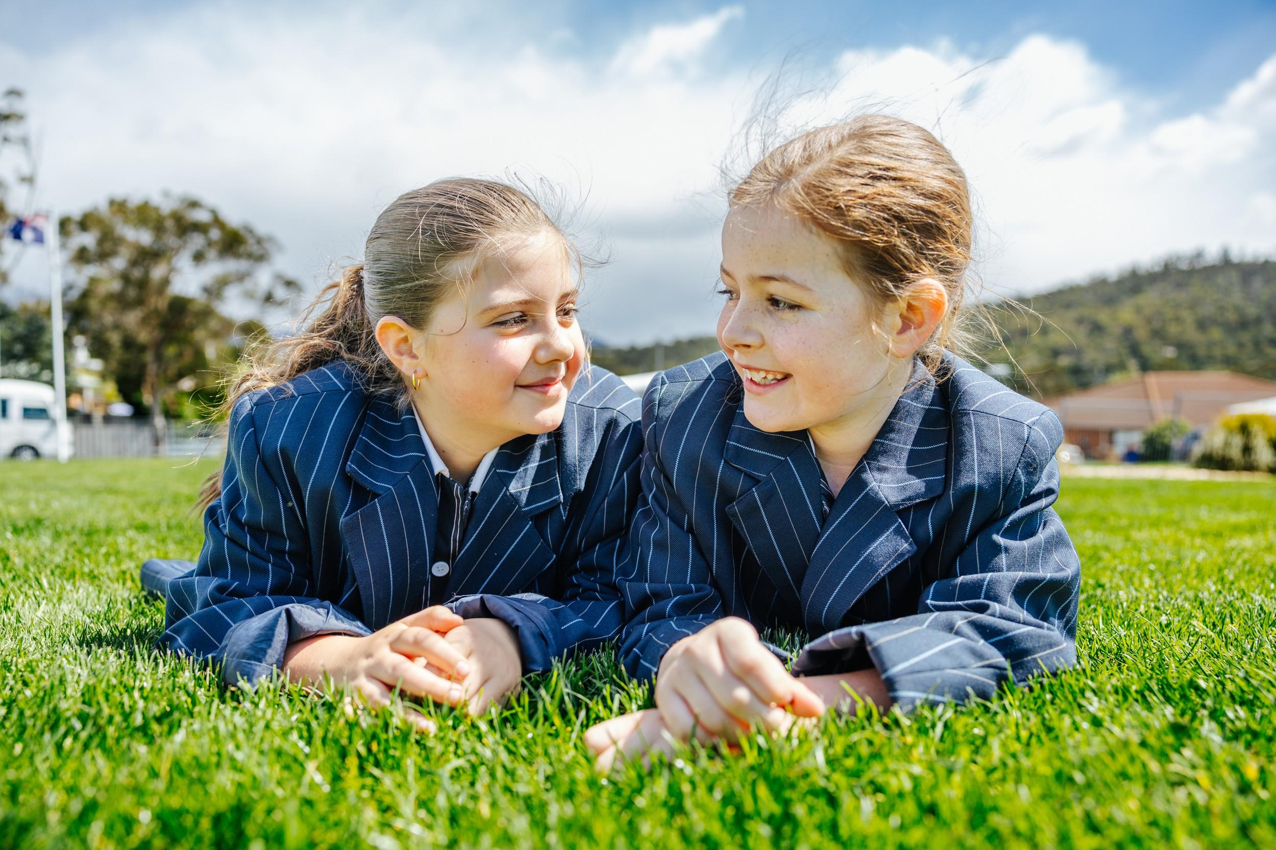 Two primary students in navy pinstripe blazers lying on the grass, smiling at each other at ACC Bairnsdale.
