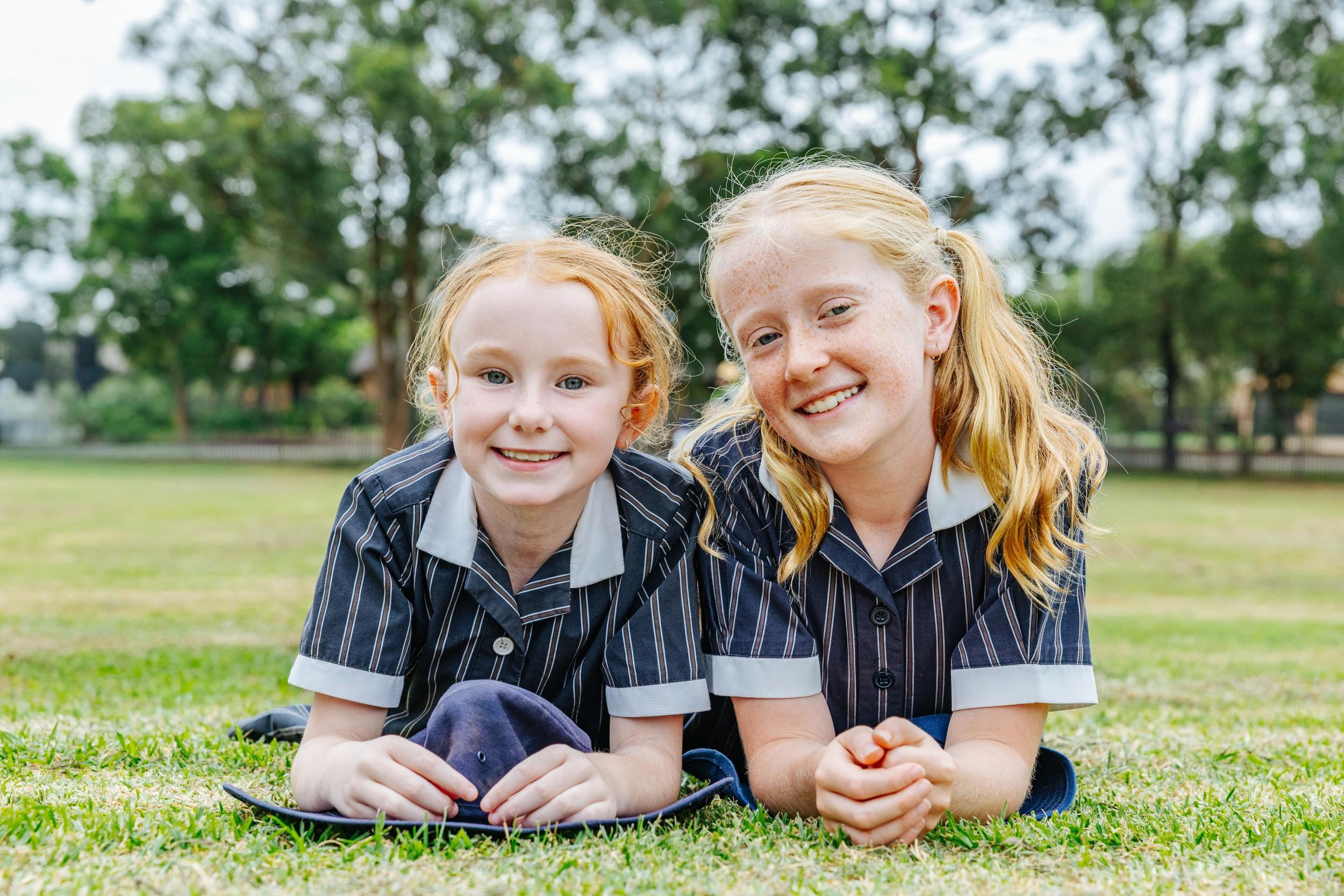 Two smiling primary students in navy pinstripe uniform lying on the grass oval at ACC Bairnsdale.