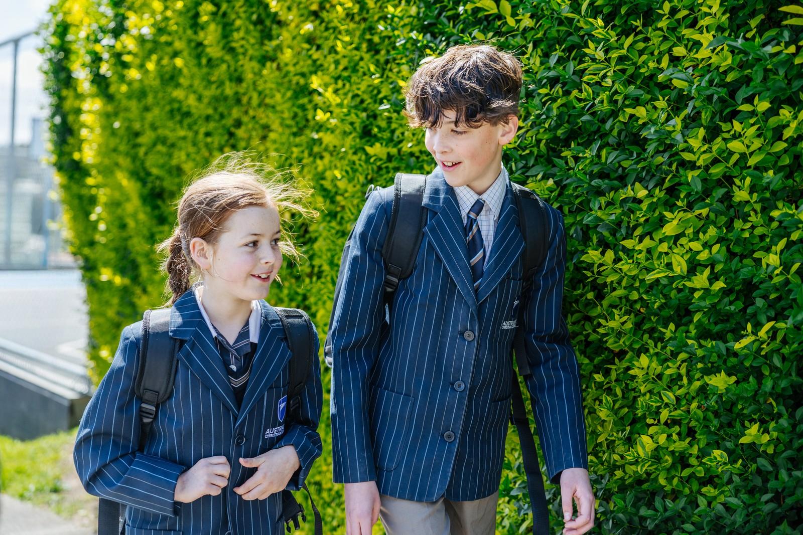 Two students in blazers and backpacks walking together along a hedge-lined path on the school grounds.