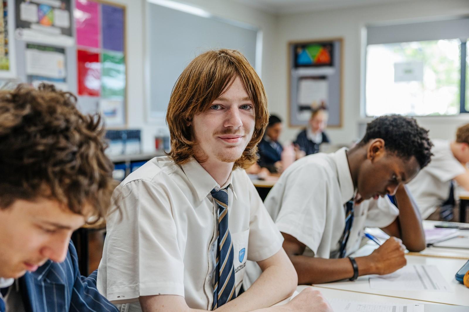 A secondary student smiling at his desk while classmates work around him at ACC Bairnsdale.
