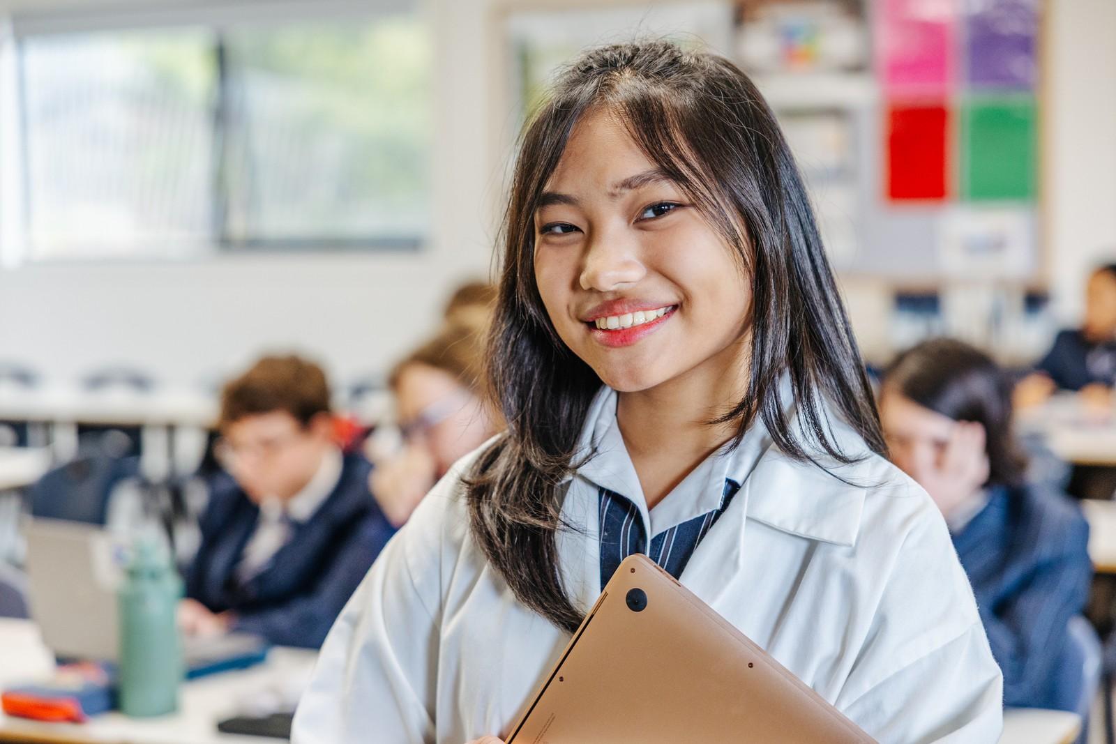 A smiling secondary student in a lab coat holding her laptop in a busy classroom.