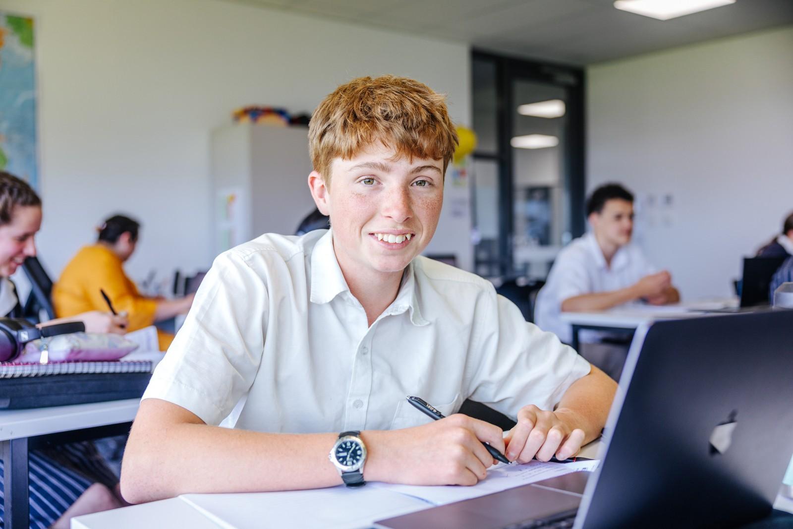 A secondary student smiling at his desk with a laptop and notes during class in Bairnsdale.