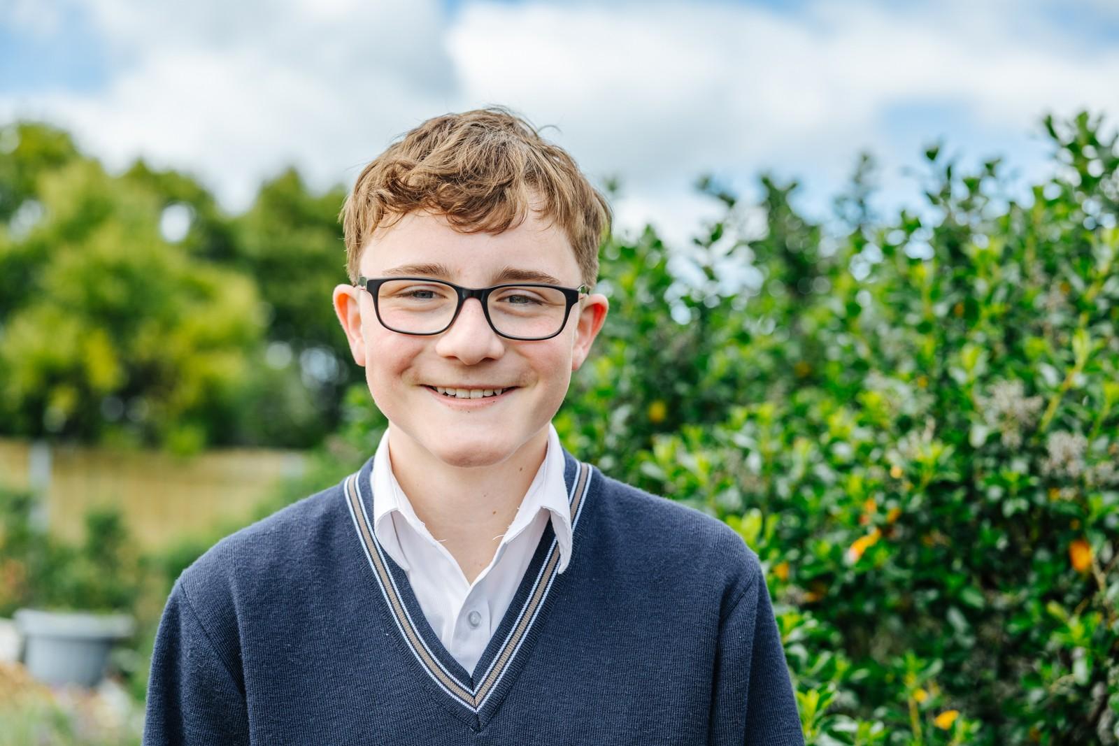 A secondary student in glasses and a navy school jumper smiling outdoors on a sunny day.