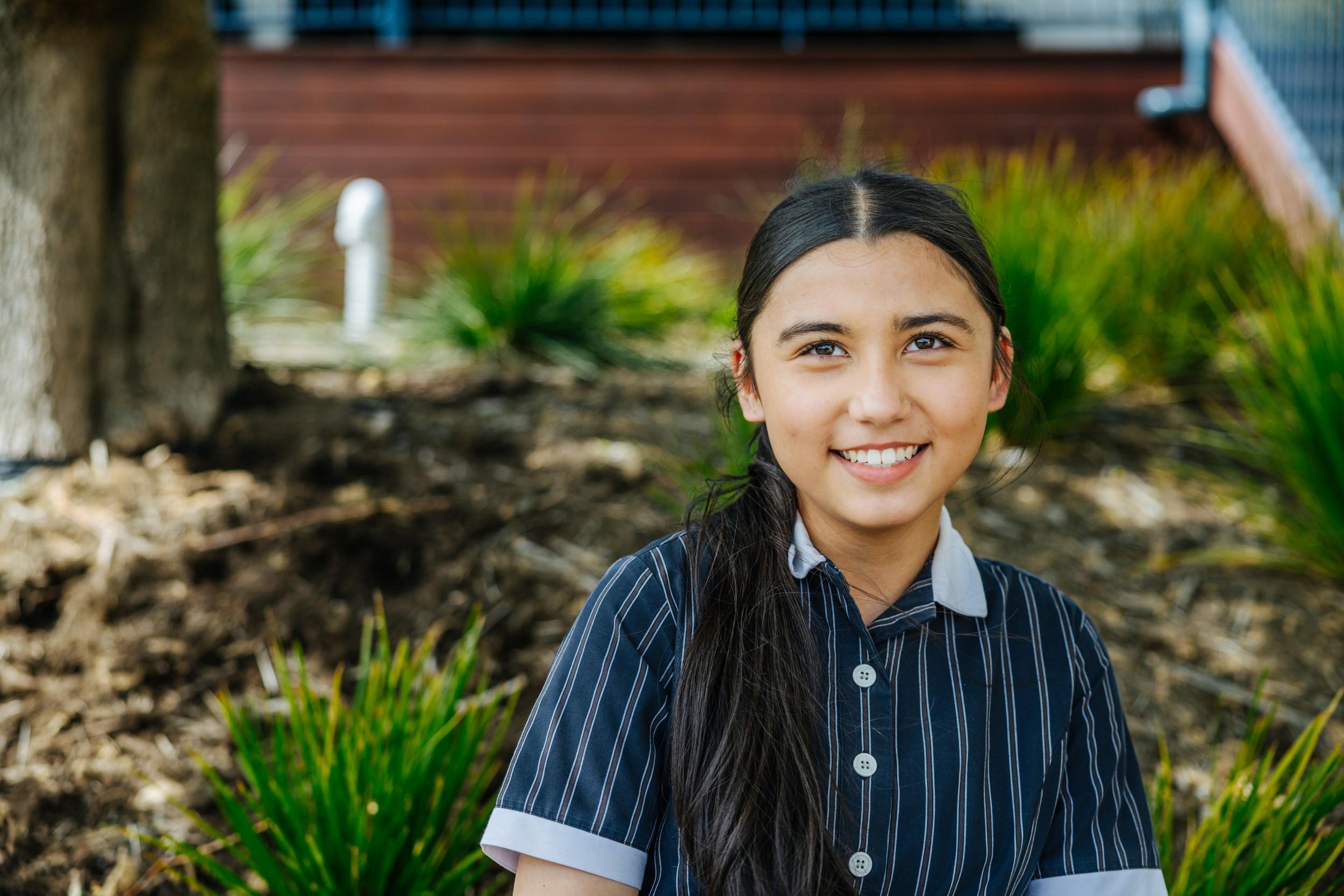 A secondary student in school uniform smiling outdoors beside the campus garden beds in Bairnsdale VIC.