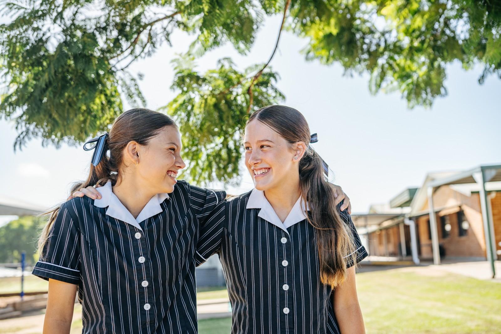 Two secondary students in striped uniforms laughing with their arms around each other beneath a shady campus tree.