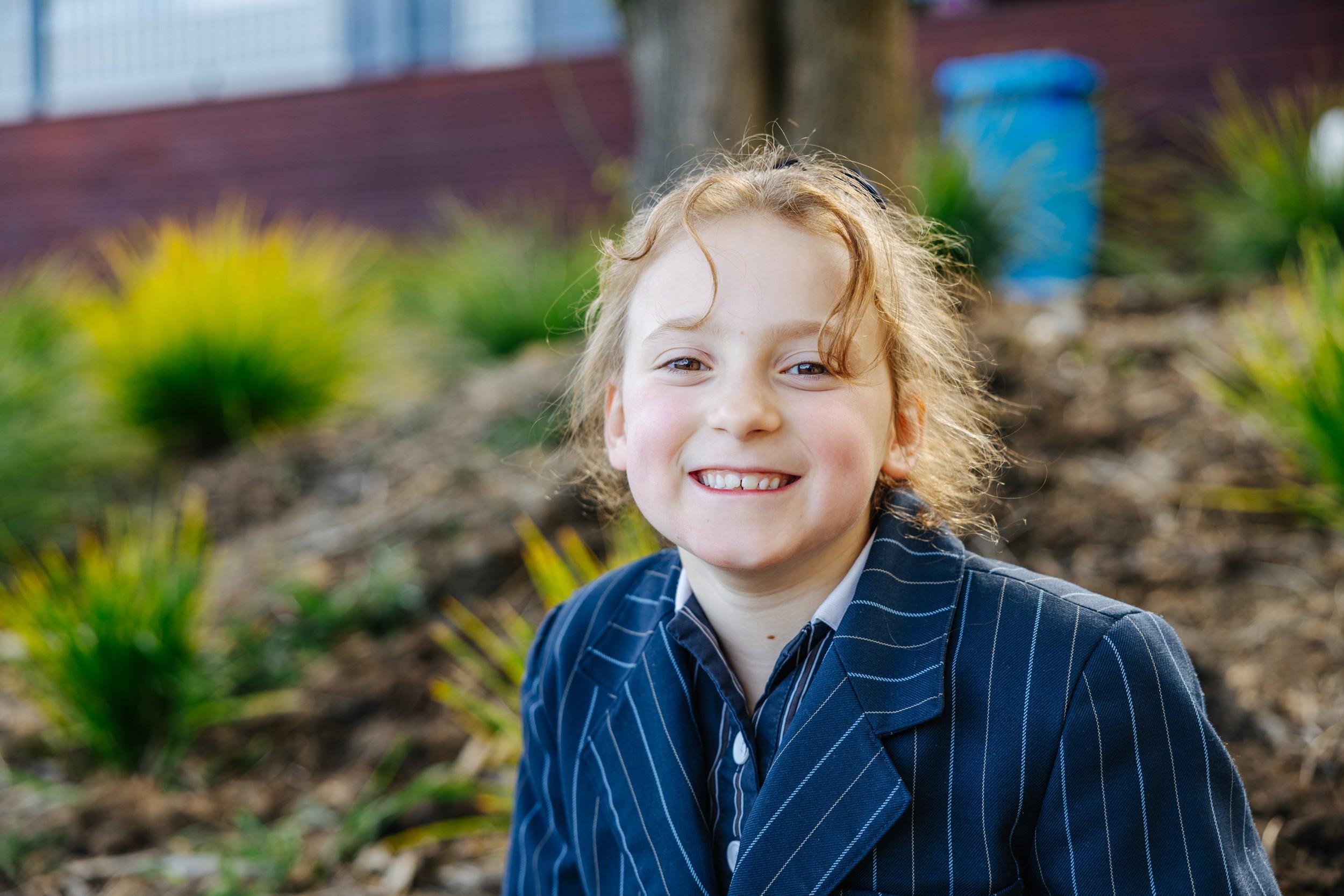 A smiling student in a navy pinstripe blazer outdoors beside the campus garden beds at ACC Bairnsdale.