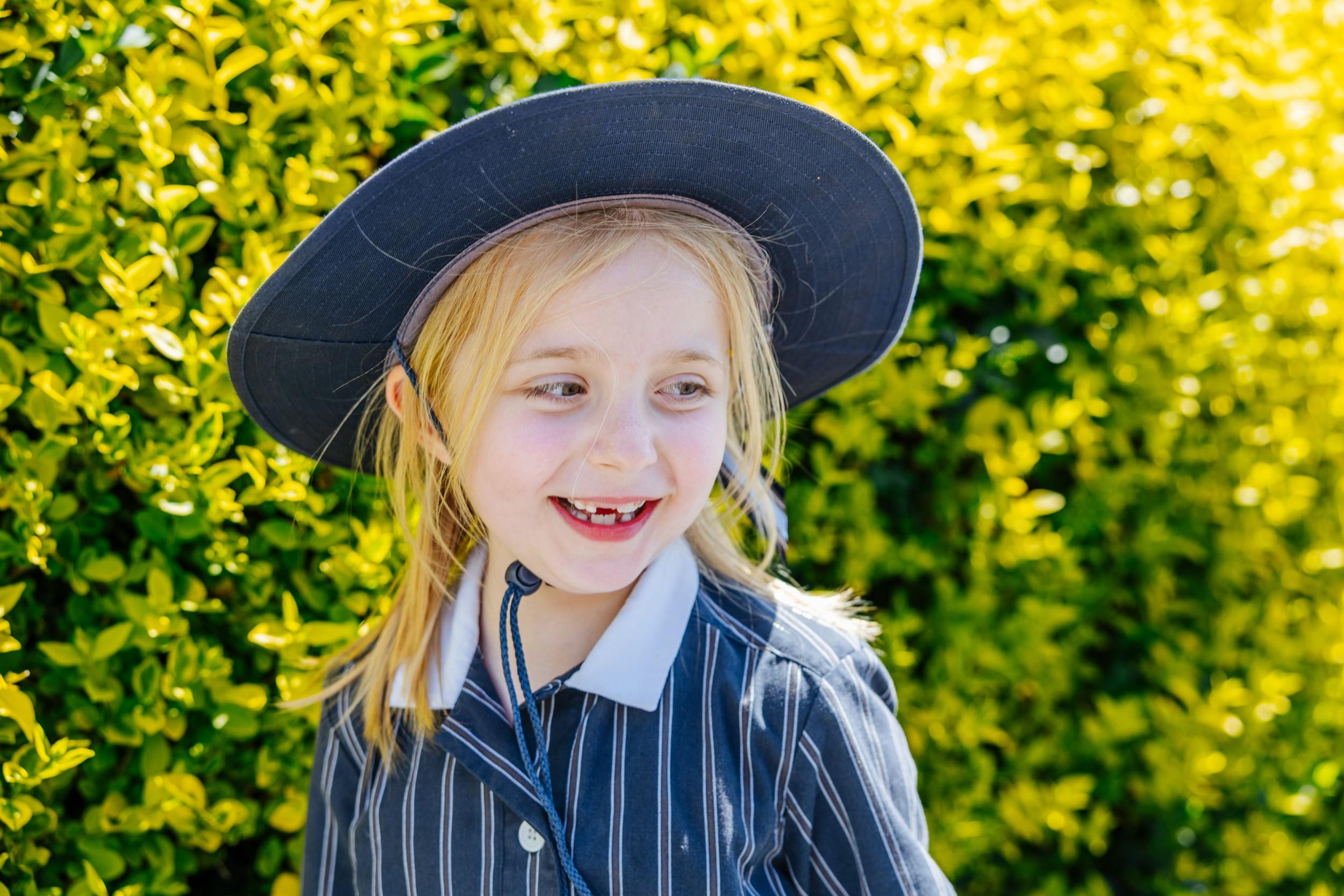 A smiling primary student in a wide-brimmed navy school hat and pinstripe uniform beside a bright green hedge.