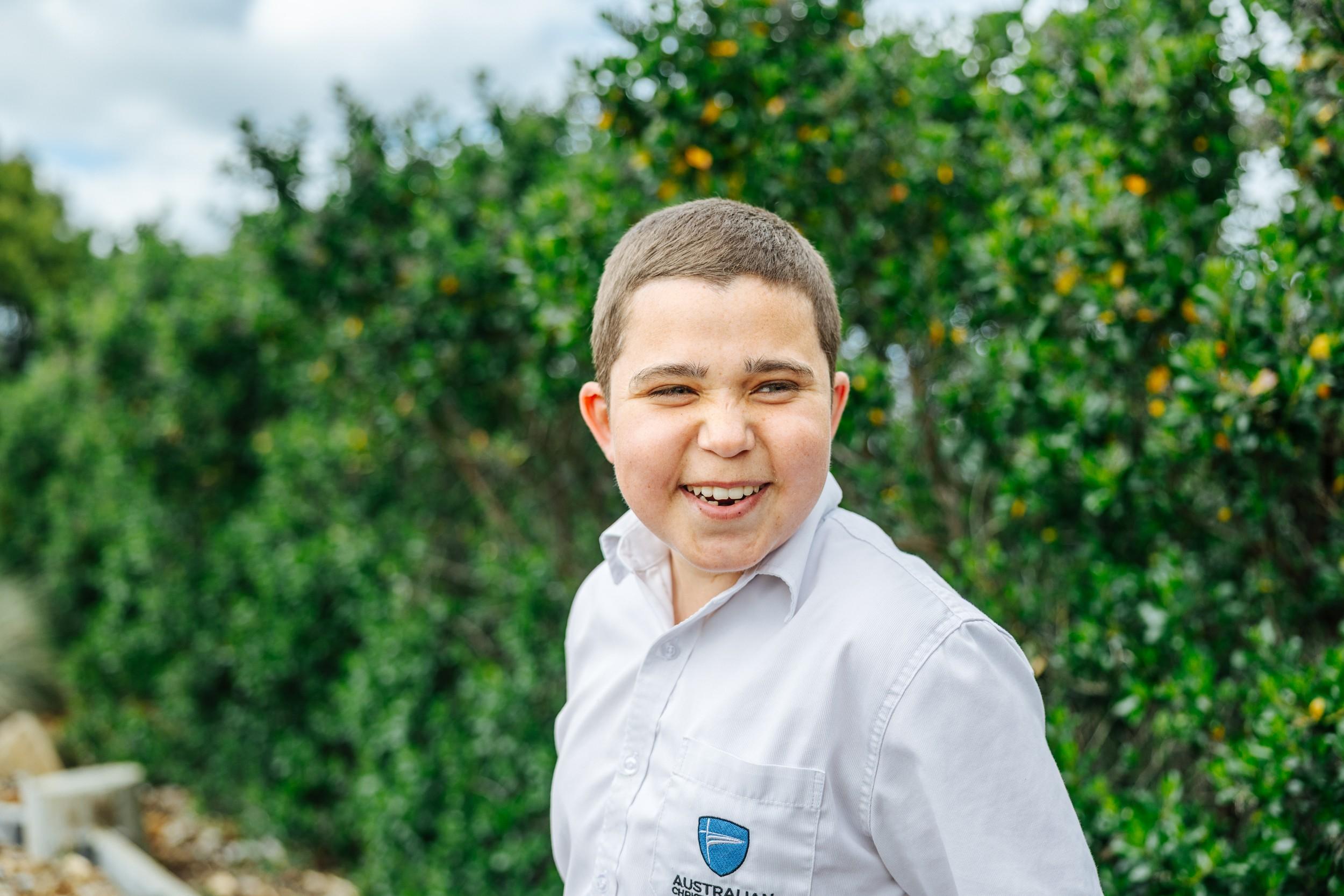 A student in an Australian Christian College shirt laughing outdoors beside a leafy hedge.