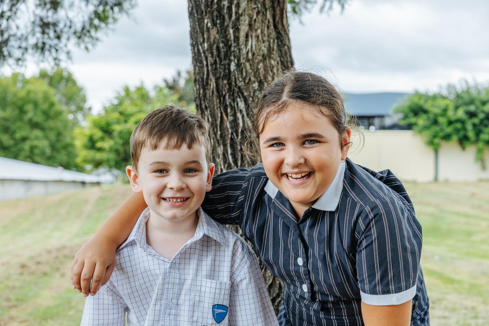 An older primary student with her arm around a younger student, both smiling outdoors under a gum tree.