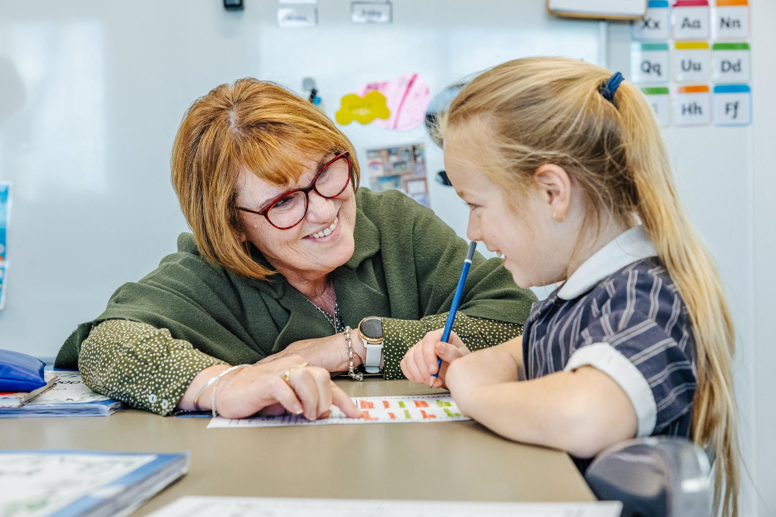 A teacher smiling beside a primary student as they work through a worksheet together at ACC Bairnsdale.