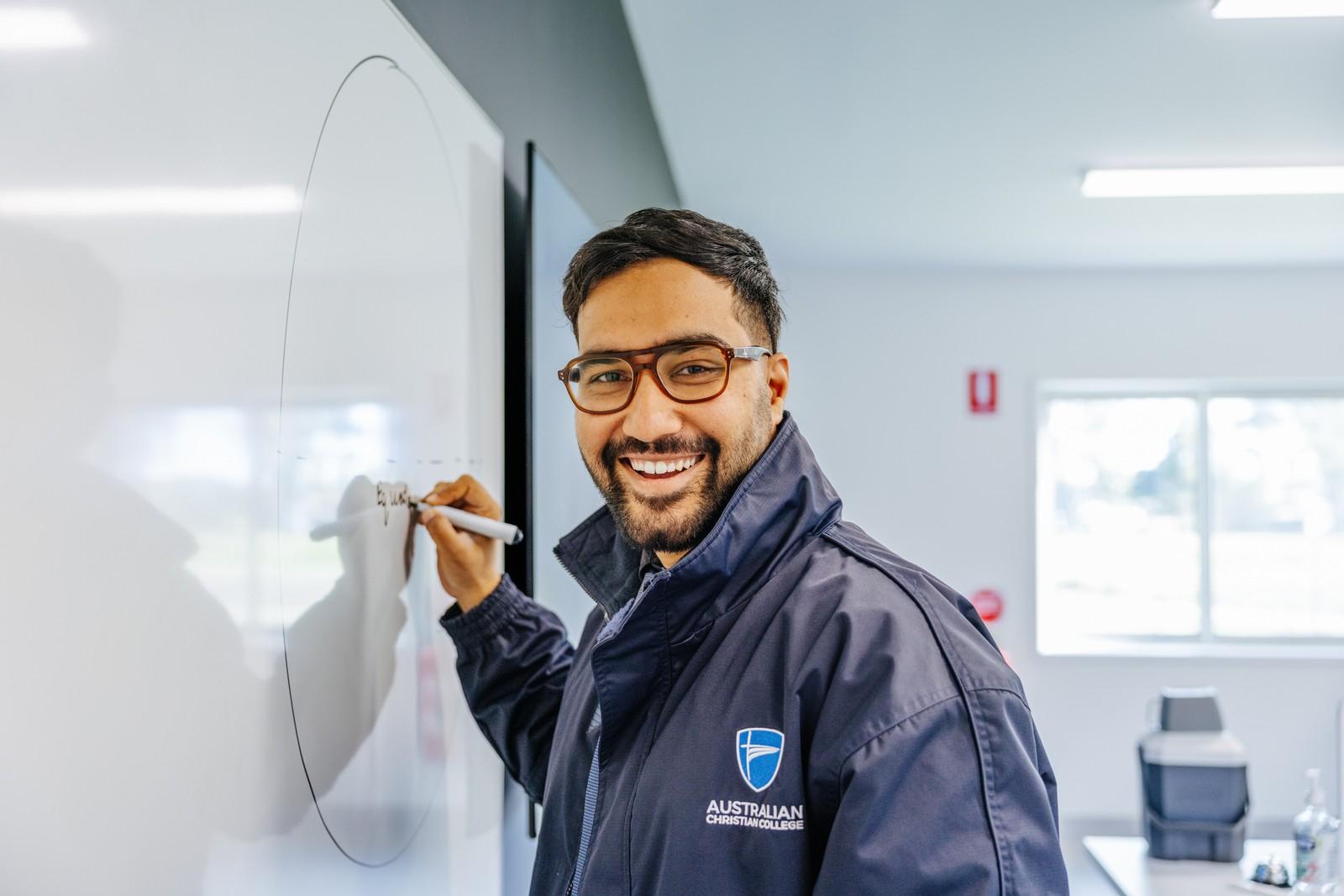 A teacher in an Australian Christian College jacket smiling while writing on a classroom whiteboard.