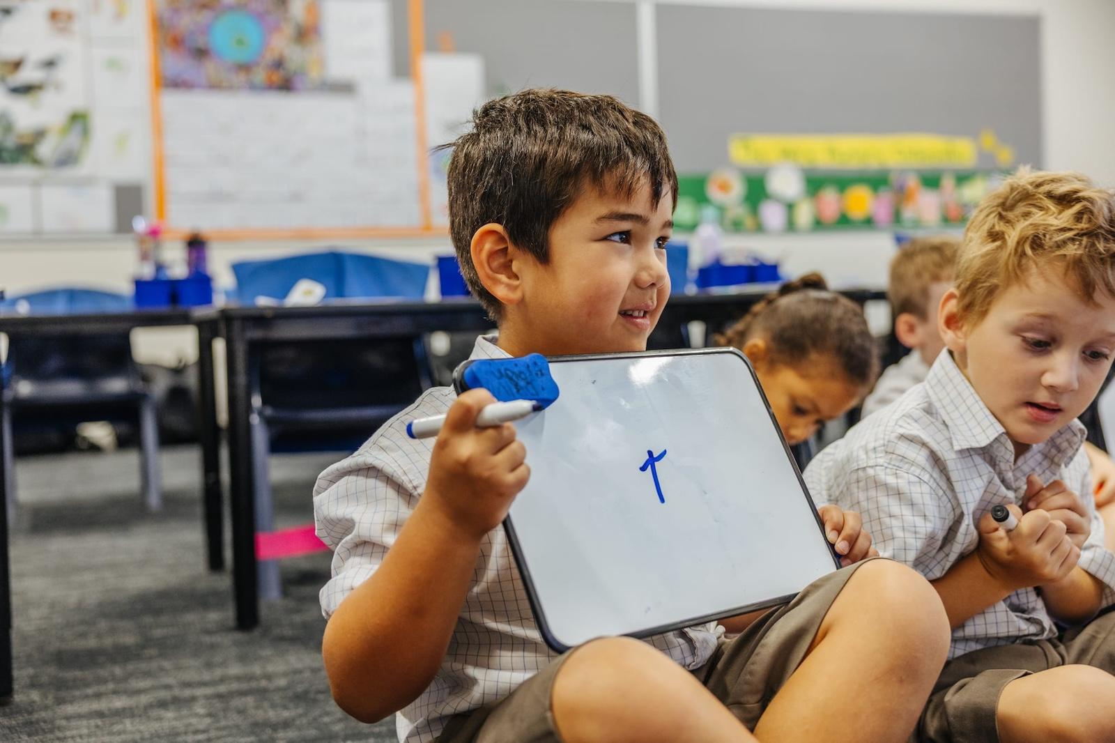A young male student holding a small whiteboard with the letter 'T' written on it, practicing Explicit Direct Instruction (EDI) in a classroom at Australian Christian College Benalla.