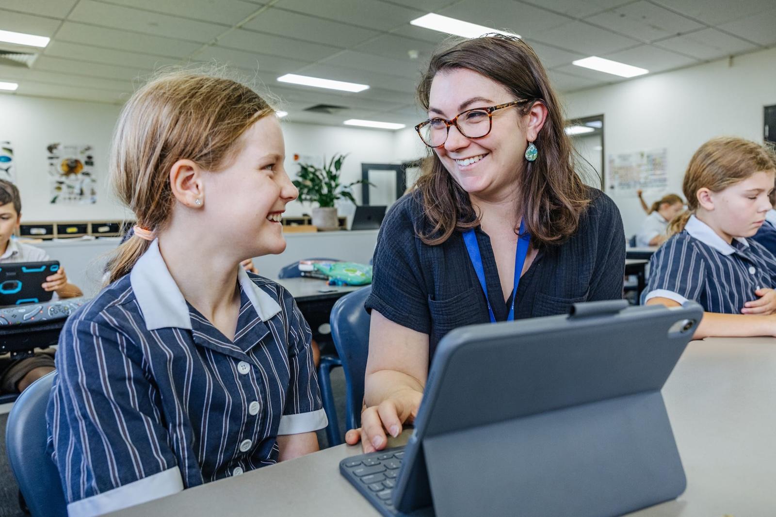A female teacher smiling and assisting a young female student with an iPad in a classroom at Australian Christian College Benalla, Victoria.