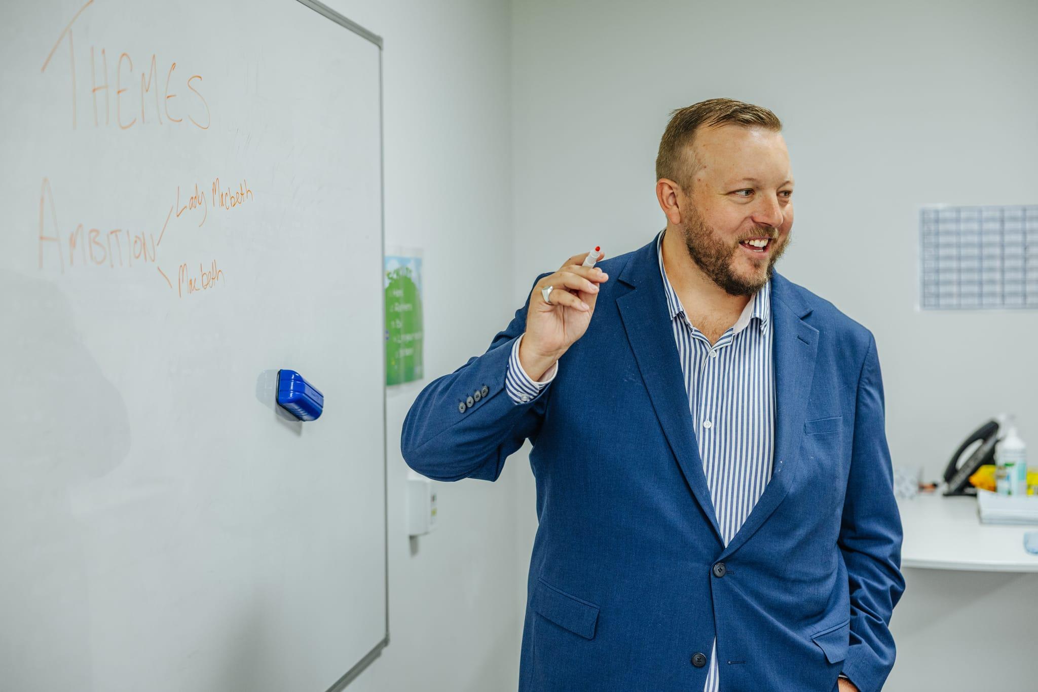 Principal of ACC Benalla, wearing a vertically striped shirt and blue blazer, smiling in front of whiteboard while teaching class.