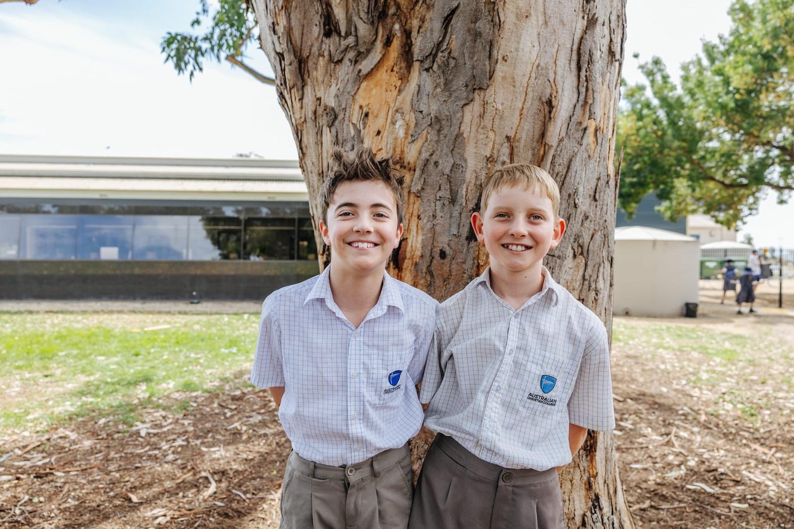 Two happy Australian Christian College Benalla students in uniform smiling by a large tree on campus, embodying the school's caring environment.