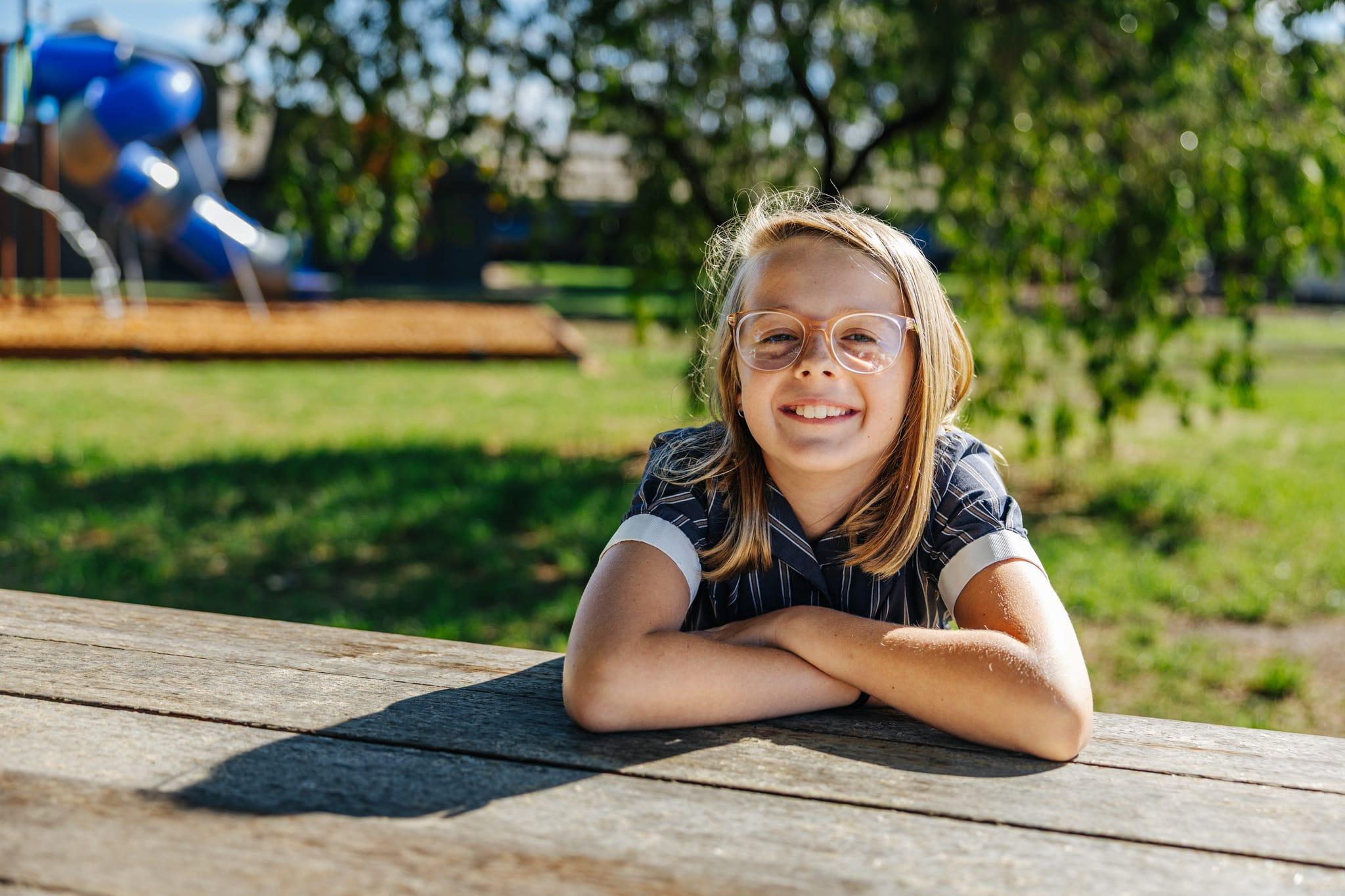 A smiling primary student wearing glasses, sitting at an outdoor wooden table, representing Christian education at ACC Benalla, Victoria.