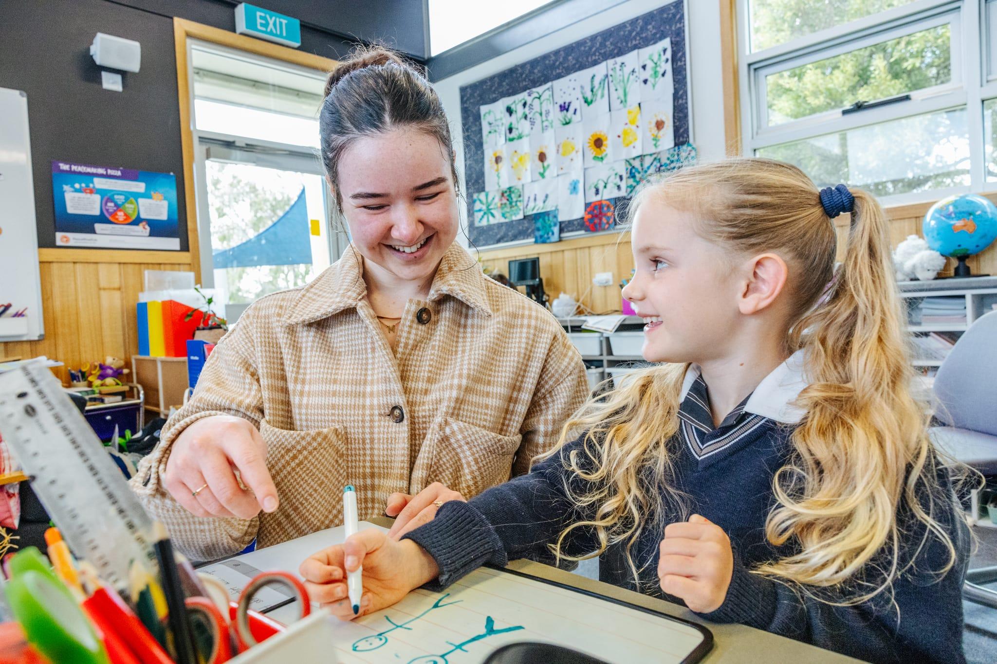A teacher smiling with a young primary student practising writing on a small whiteboard in a Somerset classroom at ACC Burnie.