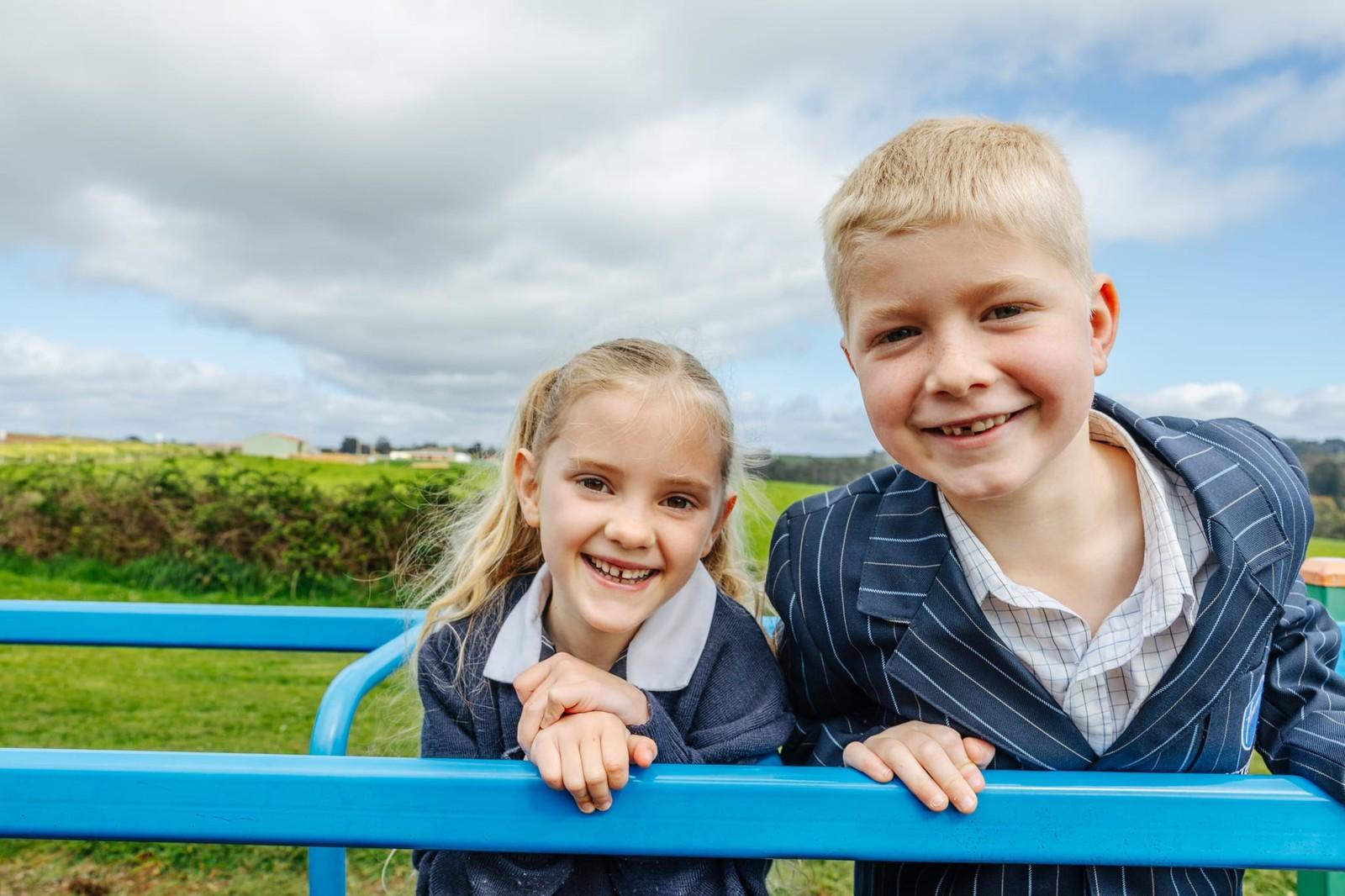 Two smiling primary students leaning on blue playground equipment with open Somerset farmland stretching behind them at ACC Burnie