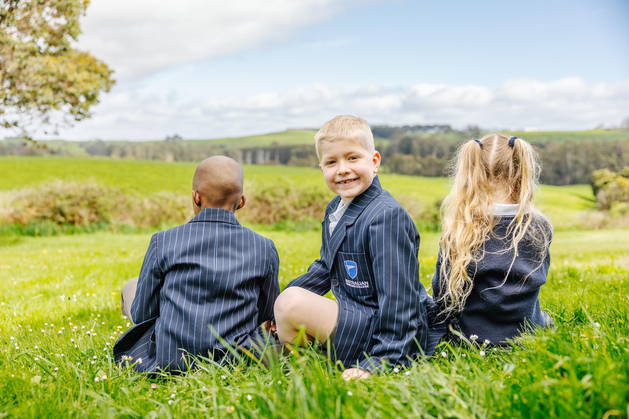 Two smiling primary school students in navy pinstripe blazers lying on green grass with Tasmanian countryside in the background.