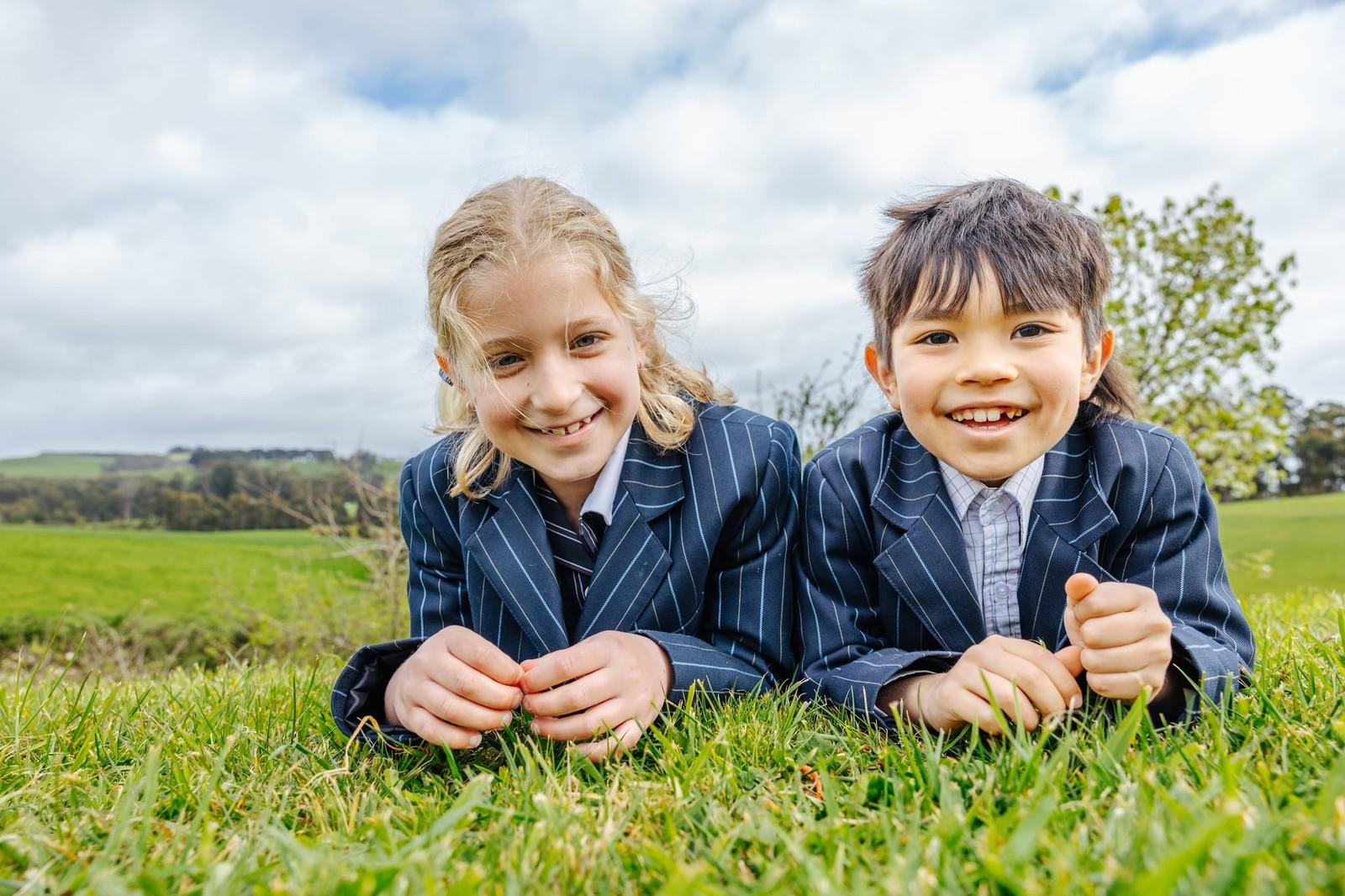 Two smiling primary school students in navy pinstripe blazers lying on green grass with Tasmanian countryside in the background.