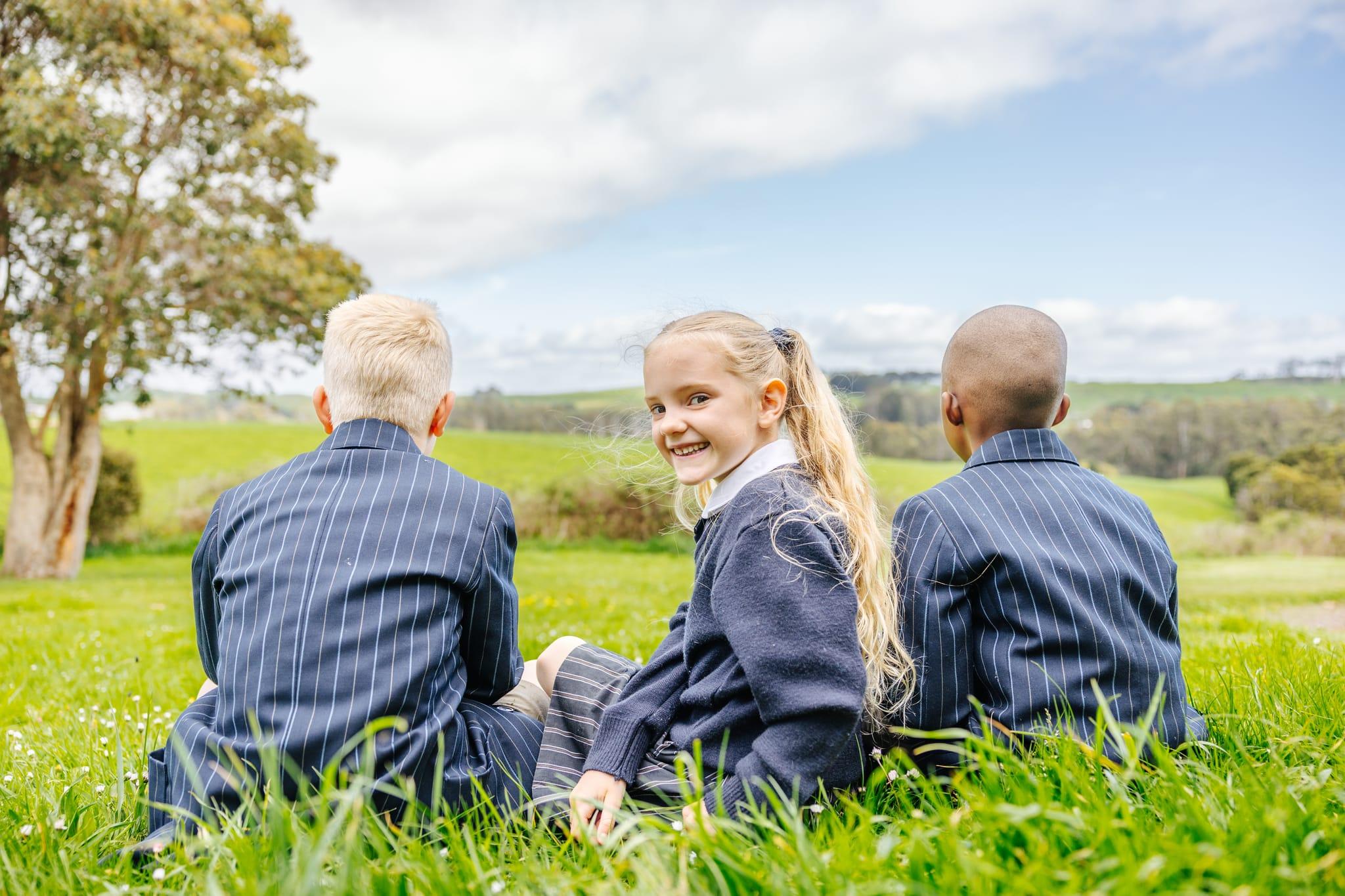 Three primary students in ACC Burnie blazers sitting together on a grassy hillside with rolling Somerset countryside behind them.