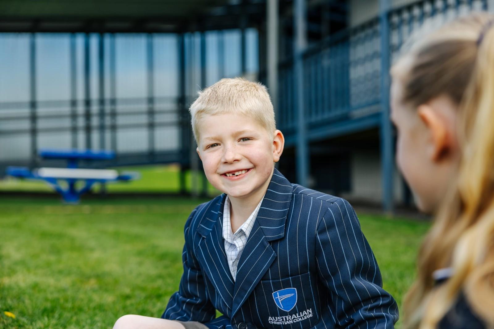 A smiling young boy in an Australian Christian College Burnie blazer seated on the grass outside school buildings in Tasmania.