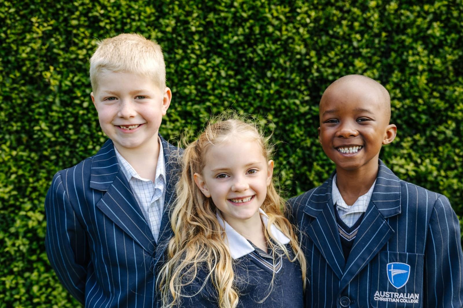 Three primary students in blazers smiling in front of a green hedge at ACC Burnie.