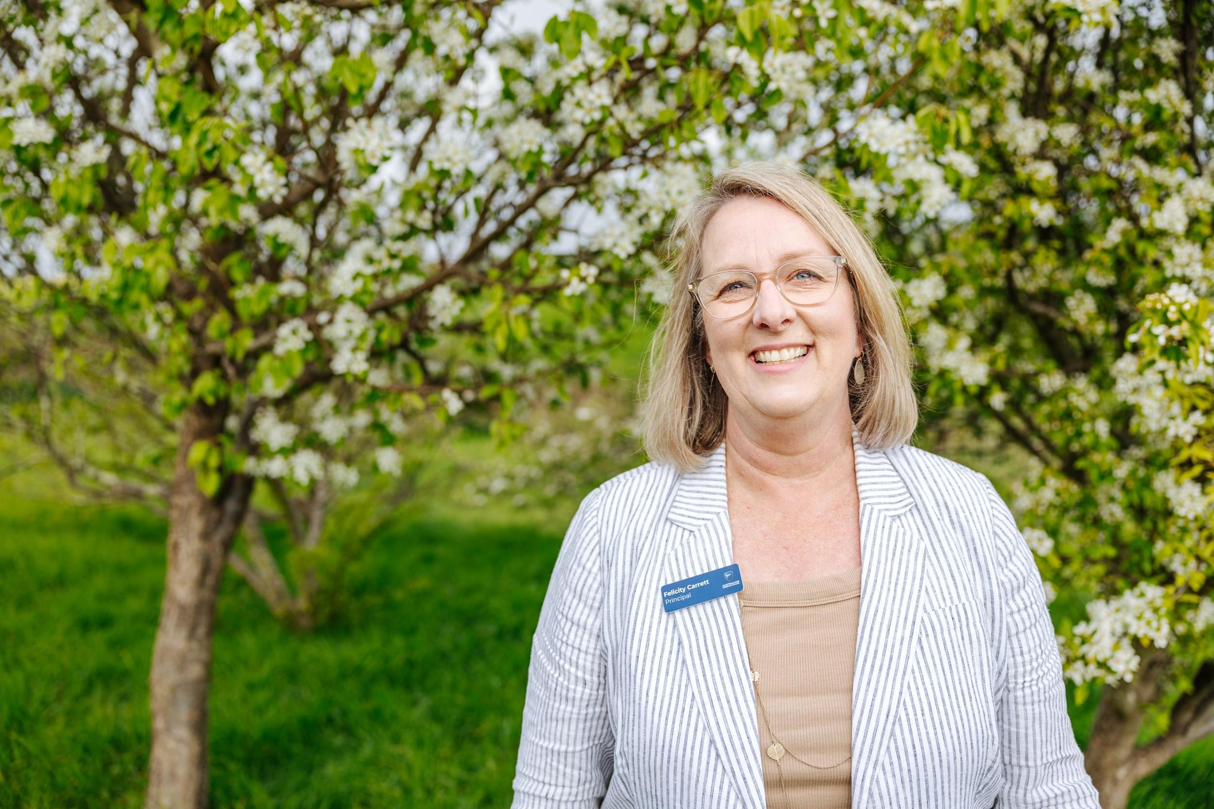 Principal Felicity Carrett smiling outdoors among flowering blossom trees at ACC Burnie in Somerset, Tasmania