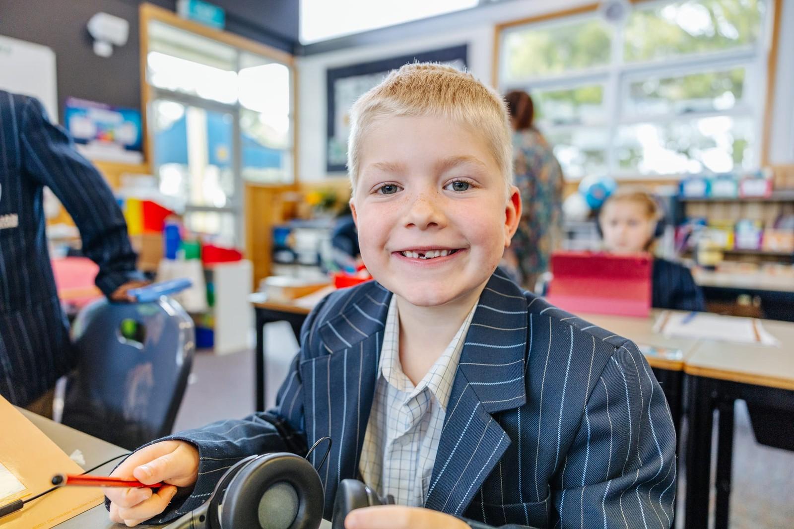 Primary boy holding headphones and a pencil, smiling confidently at his desk in an ACC Burnie classroom in Tasmania