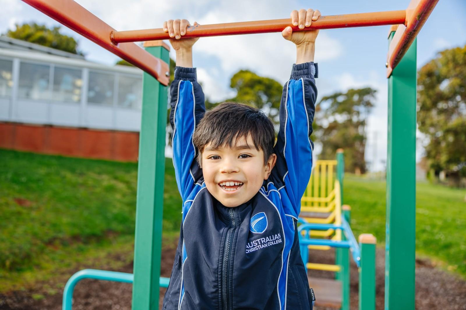Young boy in ACC uniform grinning while hanging from monkey bars on the Somerset school playground