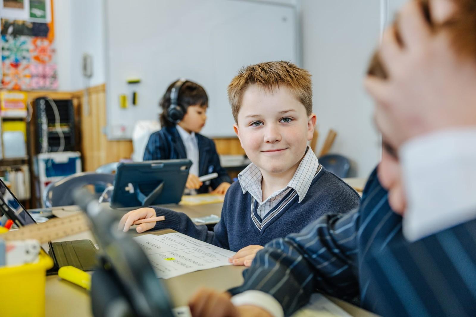 A primary school boy smiling at his desk with a worksheet and keyboard, classmates working behind him in a Somerset, Tasmania classroom.