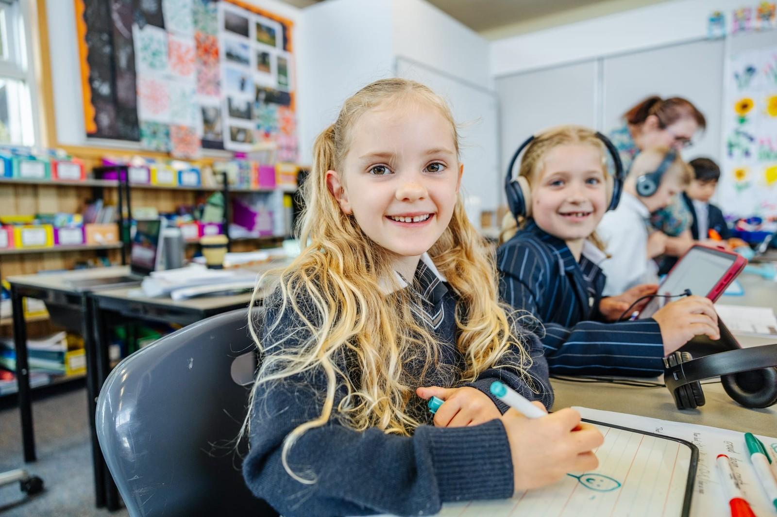 Two primary students smiling while writing on whiteboards at their desks in a bright Tasmania classroom, one wearing headphones.