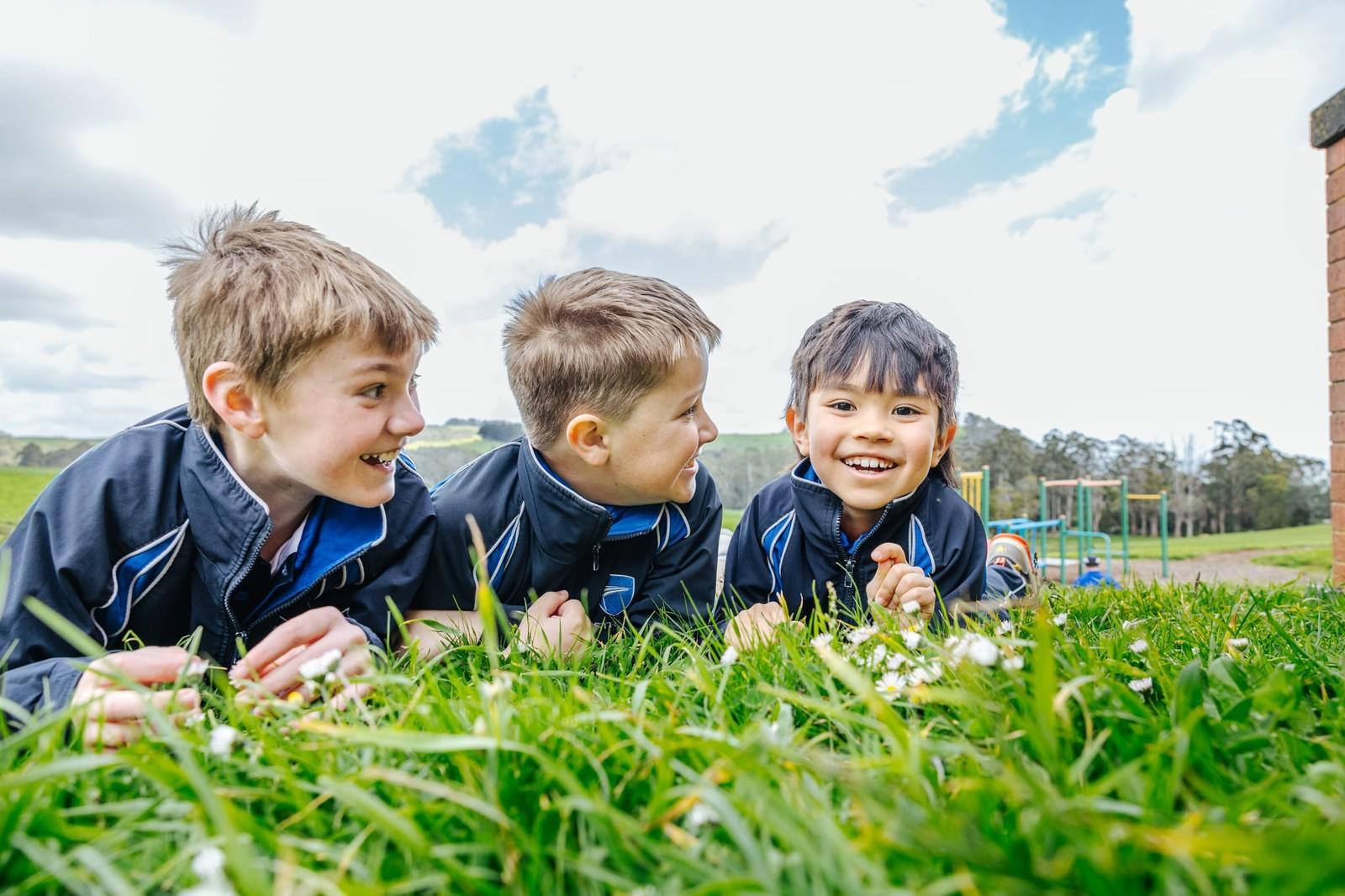 Three primary boys in sports jackets lying on grass laughing together, with a colourful playground visible in the background at ACC Burnie.