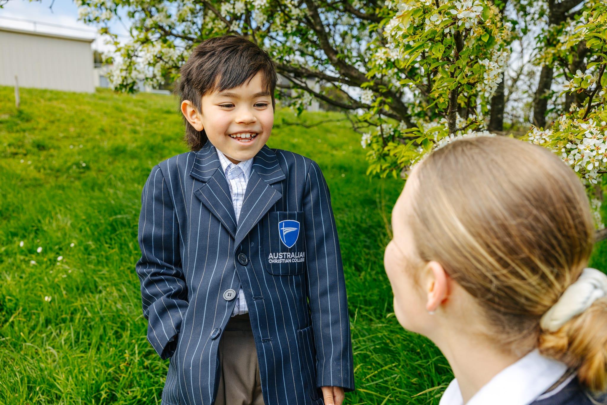 A young ACC Burnie student in a blazer smiling joyfully at a classmate under a blossoming tree on the school grounds in Somerset.