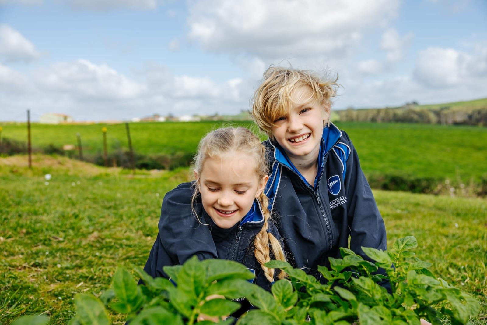 A girl and boy in ACC Burnie jackets crouching beside a garden bed, exploring plants together against a wide open Somerset landscape.