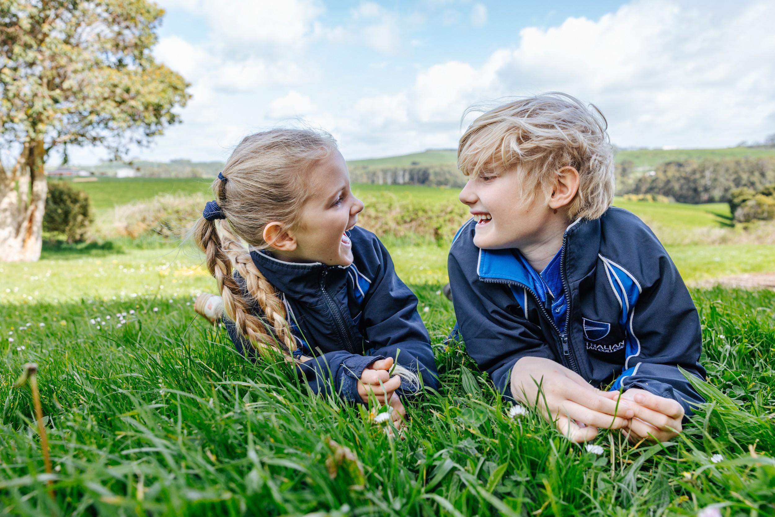 Two primary students in ACC jackets lying on green grass laughing together in the Somerset countryside at ACC Burnie.