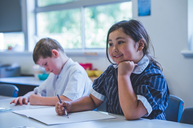 primary student resting chin on hand while completing workbook with smile