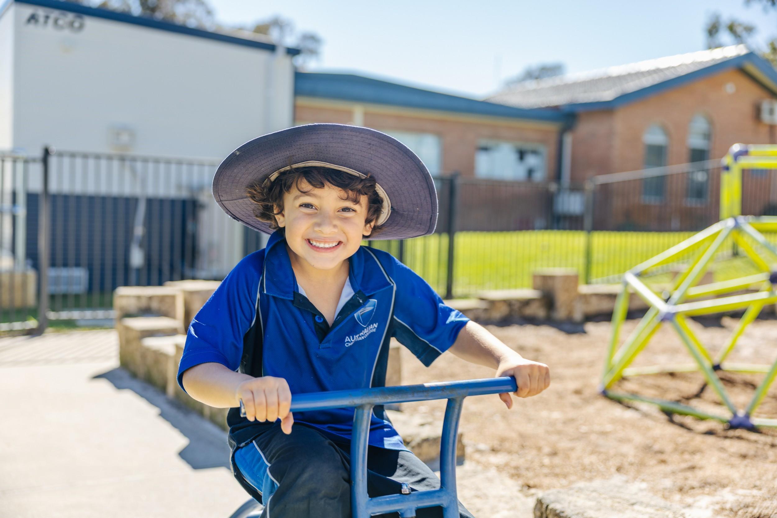 Smiling Kindergarten student in a sun hat plays outdoors on the playground at ACC Darling Downs.