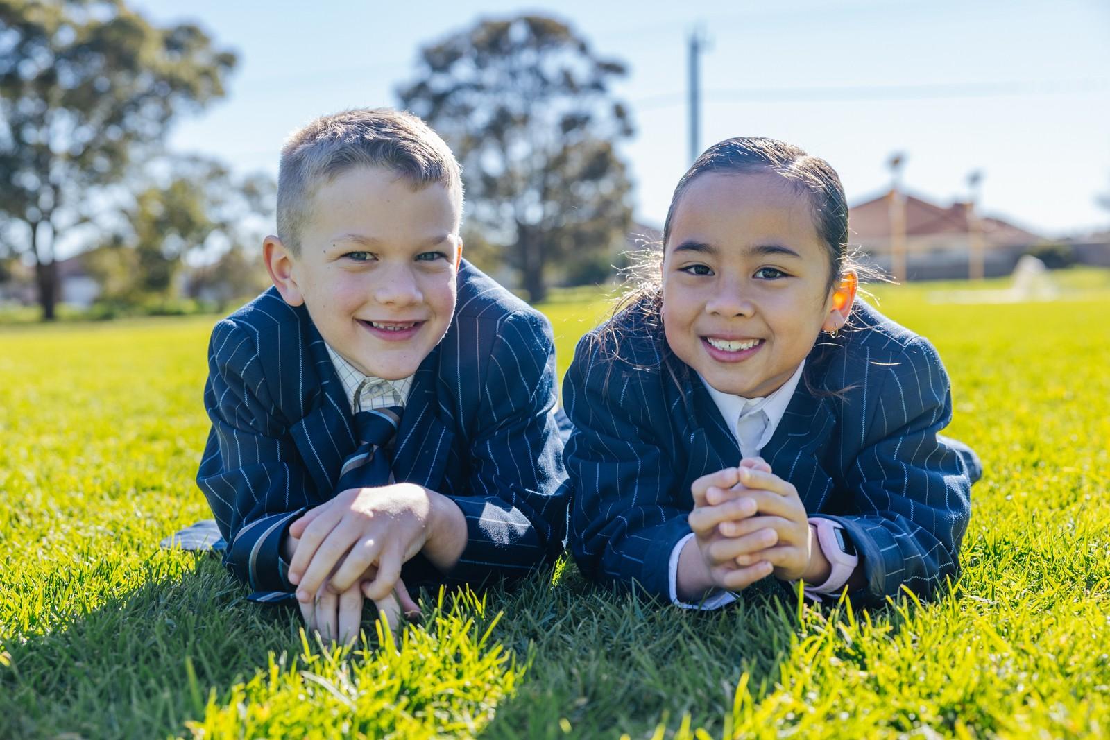 Two primary students in uniform smiling while lying on the grass at Darling Downs
