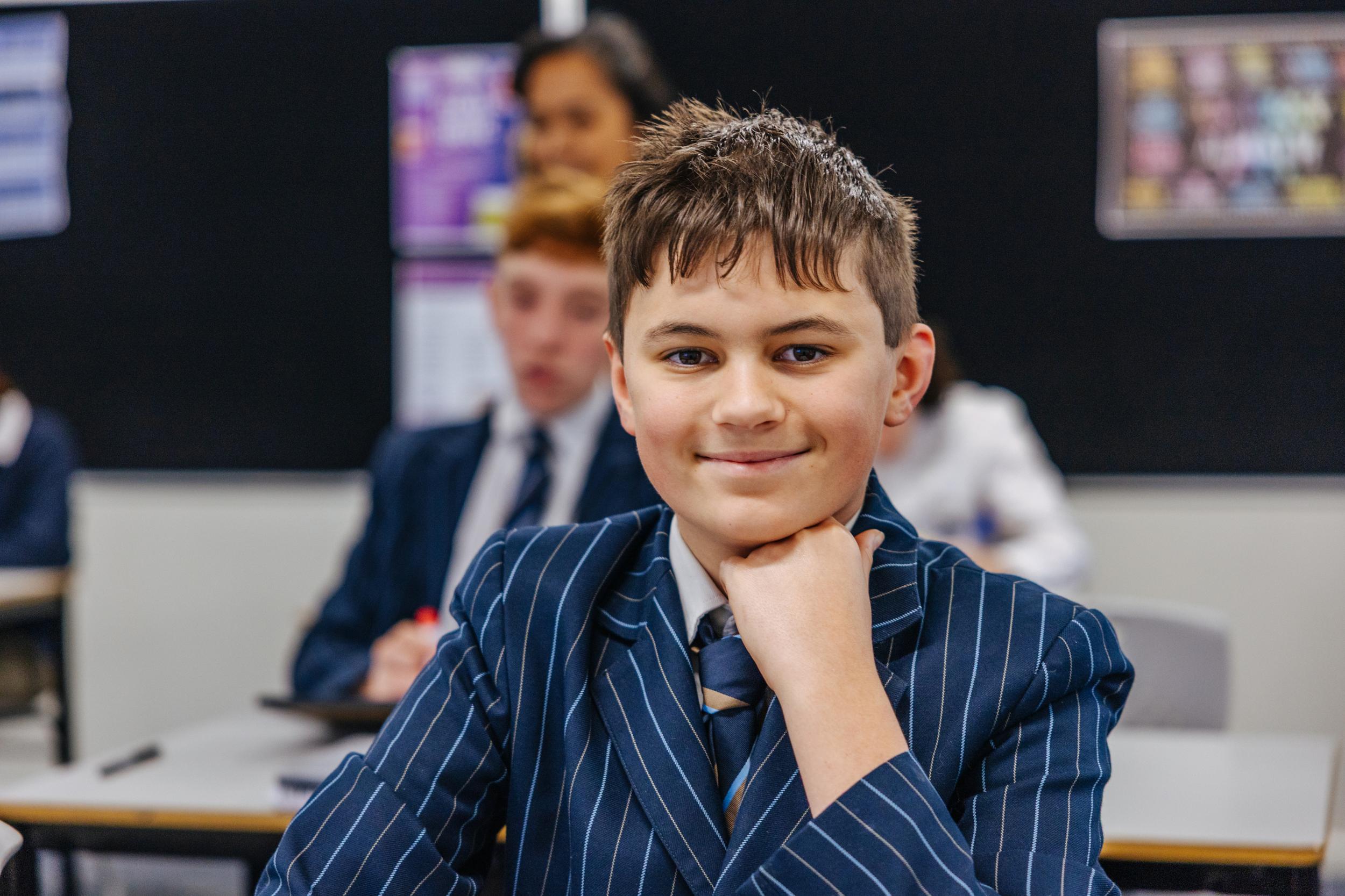 A secondary student smiles at her desk while a classmate studies nearby, reflecting the supportive Darling Downs community for teens.