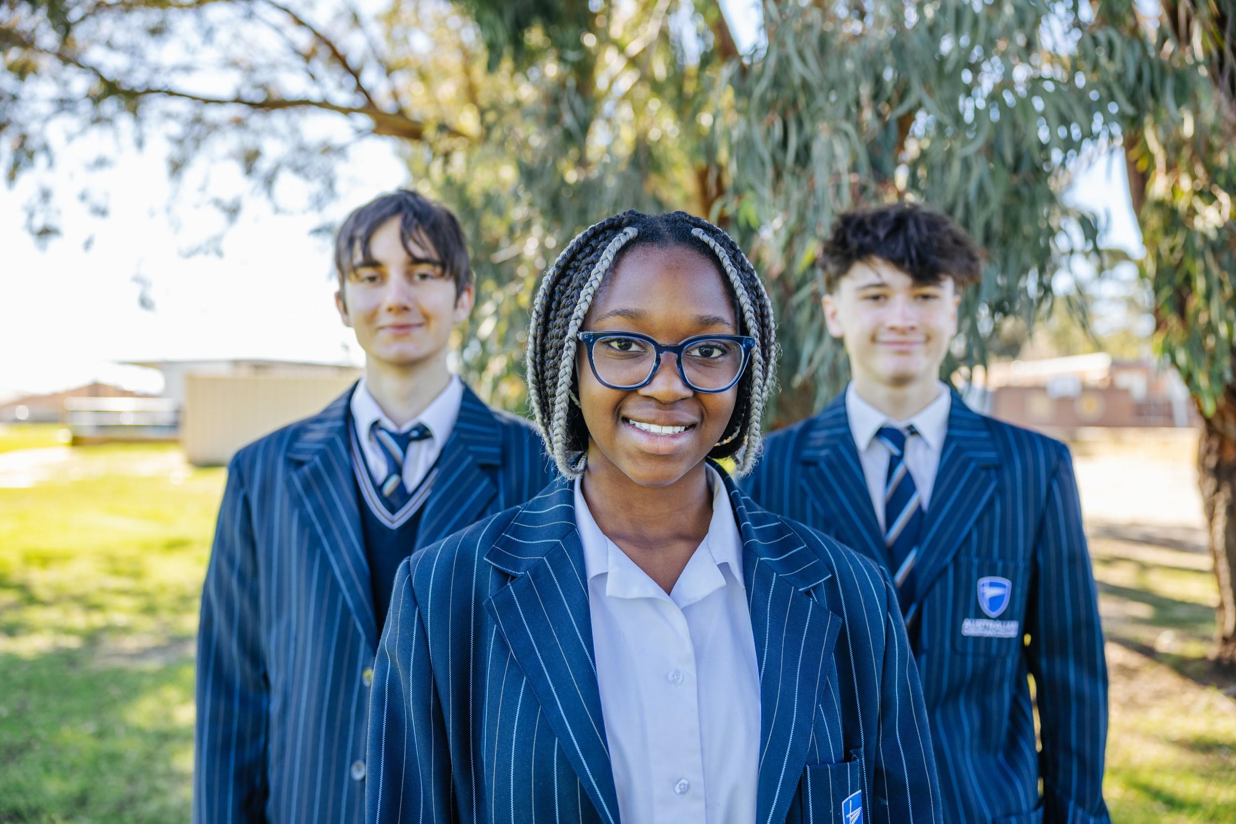 Three smiling secondary students in blazers standing outdoors among gum trees at ACC Darling Downs