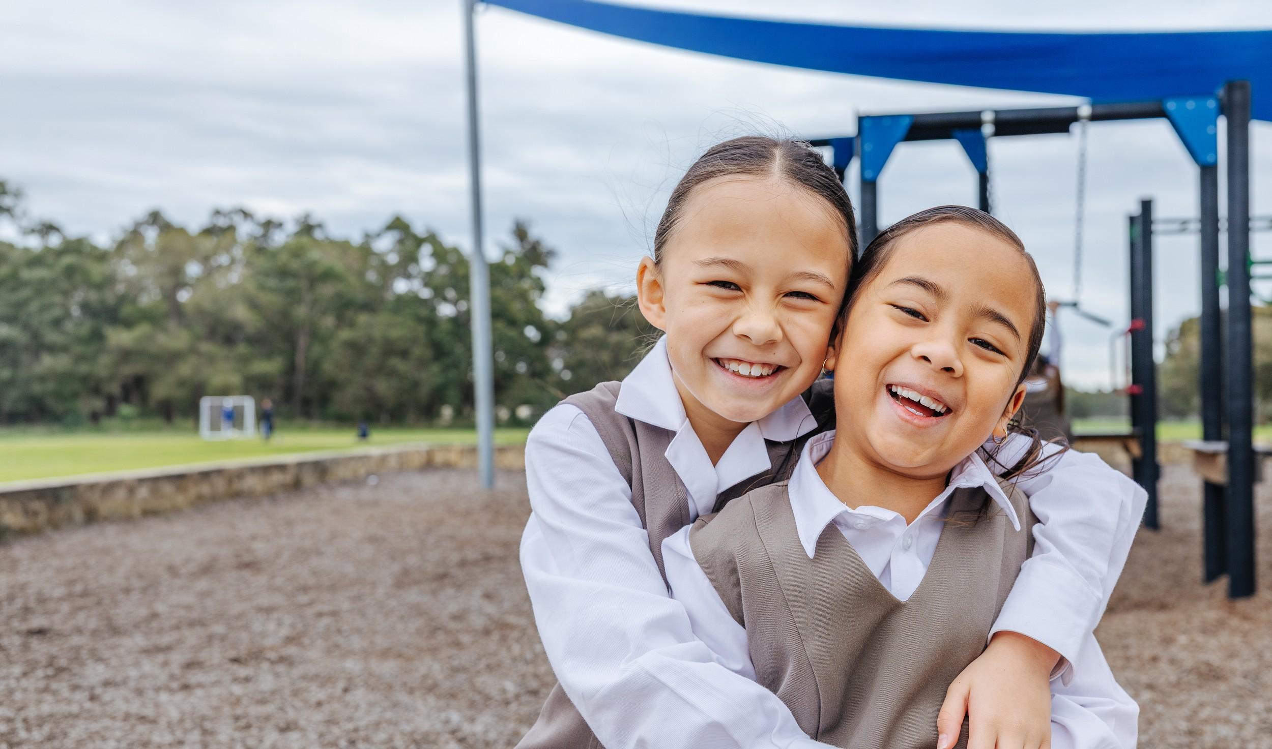 Two primary students hug and smile by the playground at ACC Darling Downs, reflecting a well loved school community.