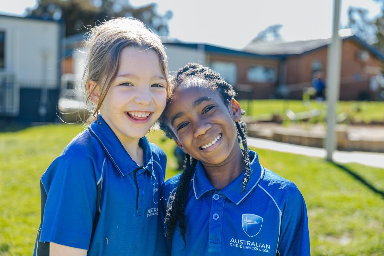 Two students lean together and smile outdoors, showing the everyday friendship and care of a well loved school.