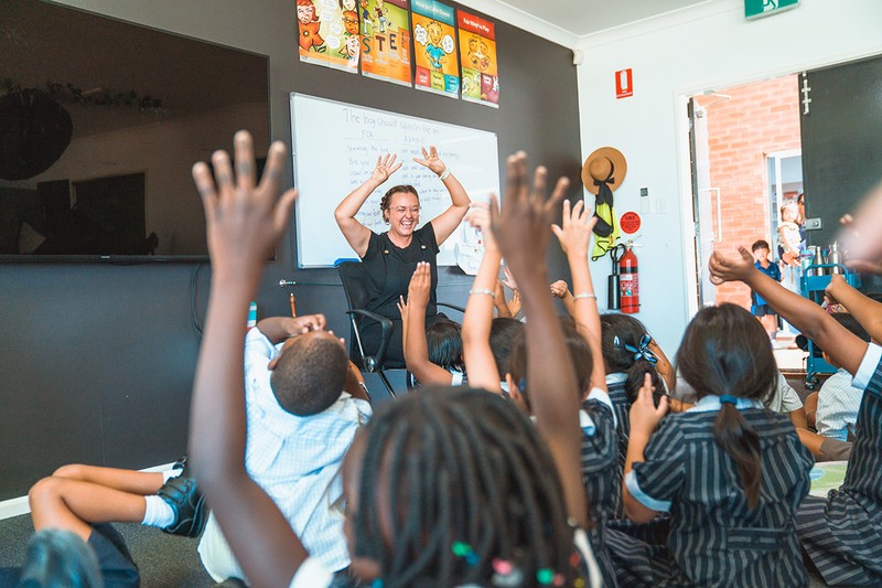 Classroom of kindergarten students with hands raised looking at smiling teacher