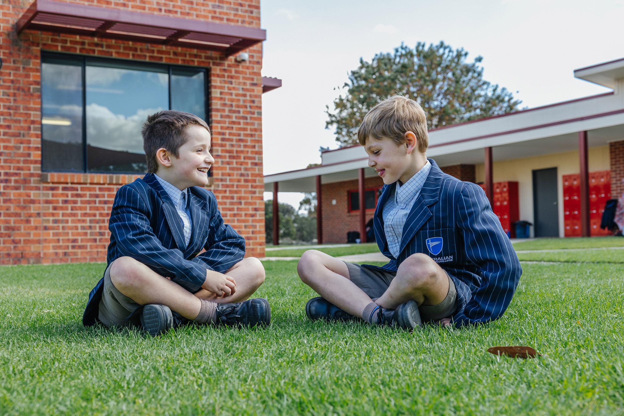 Two students sit on the lawn in school uniform outside ACC Echuca’s red-brick classrooms, representing a safe and structured environment shaped by clear school policies.