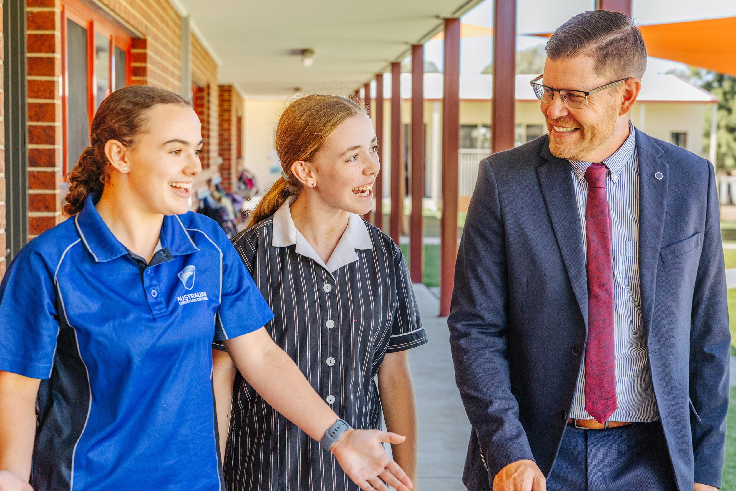 ACC Echuca principal walks alongside two smiling secondary students during class changeover, modelling approachability and servant-hearted leadership in a vibrant school setting.