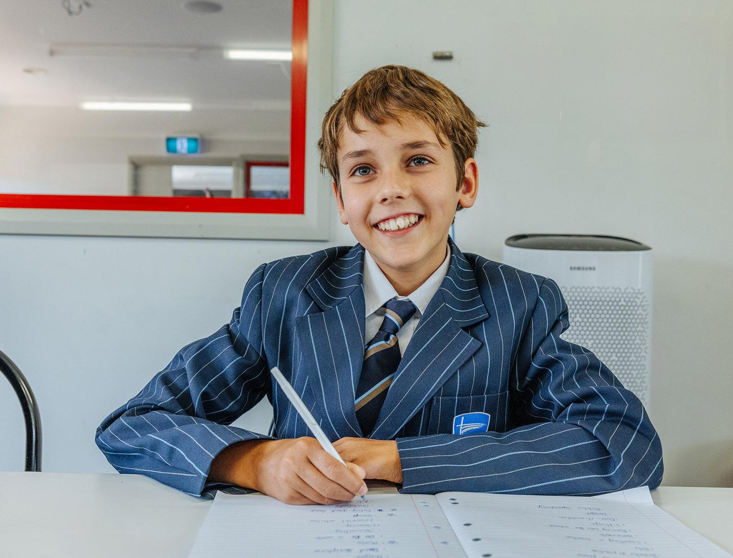 Smiling primary school boy in pinstripe uniform writing in a nuturing Christian classroom at ACC Echuca.