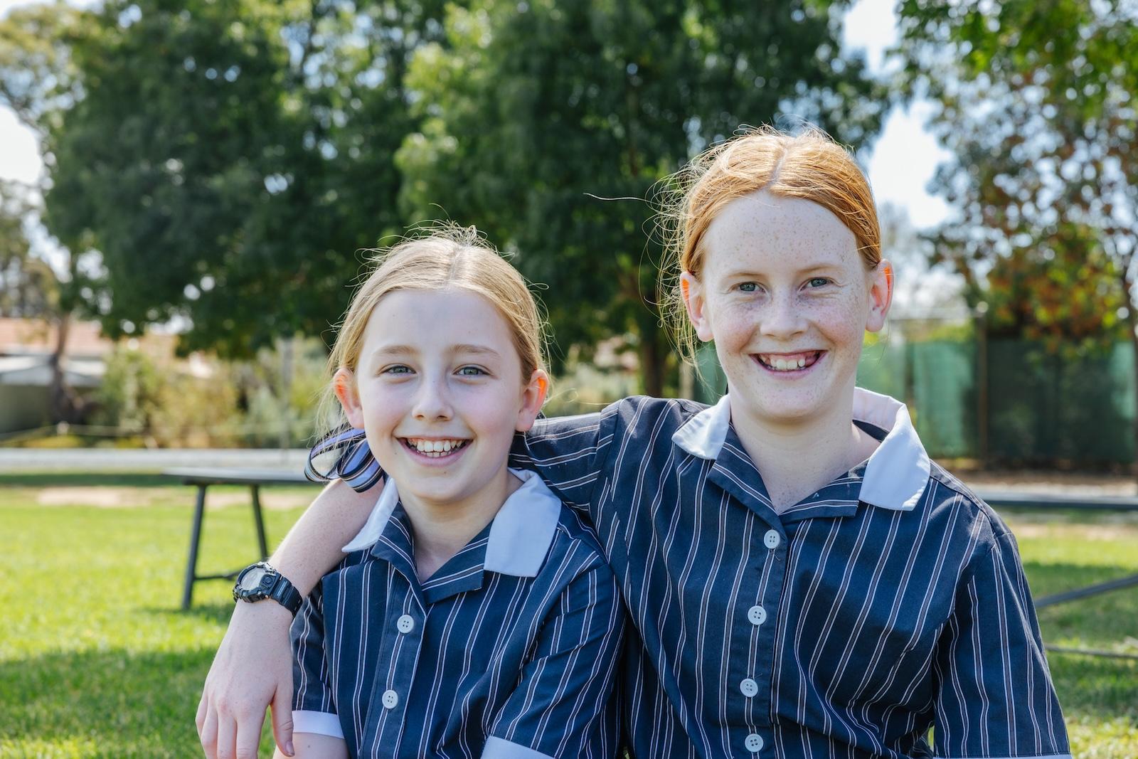 Two smiling primary school girls arm in arm on the school lawn at ACC Echuca, representing a supportive and Christ-centred Christian school community.